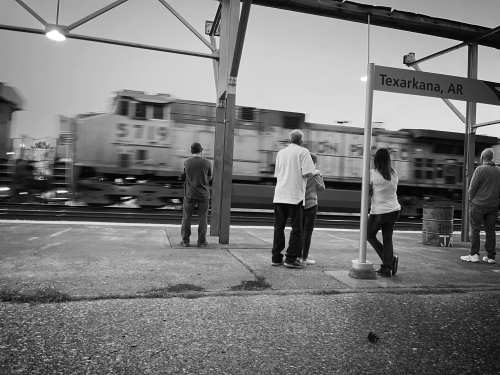 A black and white image of people waiting at a train station as a train passes by in the background.