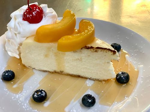 A slice of cheesecake topped with peach slices, whipped cream, a cherry, and blueberries, drizzled with syrup.