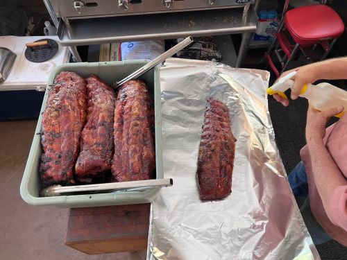 A tray of smoked ribs next to a single rib on foil, with someone spraying the ribs in a food preparation area.