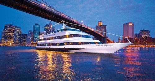 A white cruise ship named "Spirit of New Jersey" sails under a bridge with a city skyline at dusk.