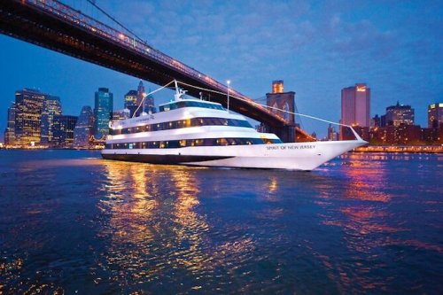 A cruise ship named "Spirit of New Jersey" glides on water beneath a city skyline at dusk.