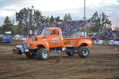 An orange vintage truck named "Agent Orange" competes in a dirt arena, with a crowd in the background.