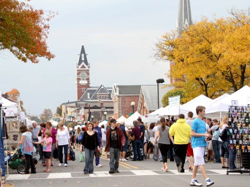 A bustling outdoor market with people browsing tents, colorful autumn trees, and a clock tower in the background.
