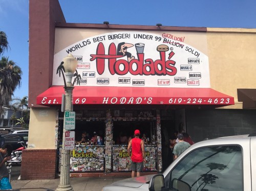A colorful restaurant exterior with a sign reading "Hodad's" and people dining outside in a lively atmosphere.