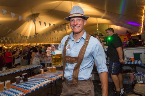 A man in traditional attire holds a large beer mug at a festive outdoor event with people and decorations in the background.