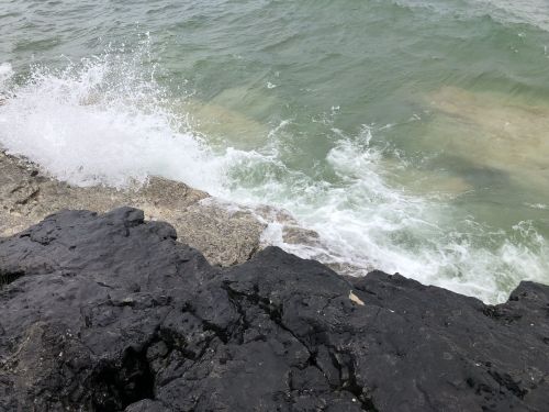 Waves crashing against dark rocky shore with clear green water in the background.