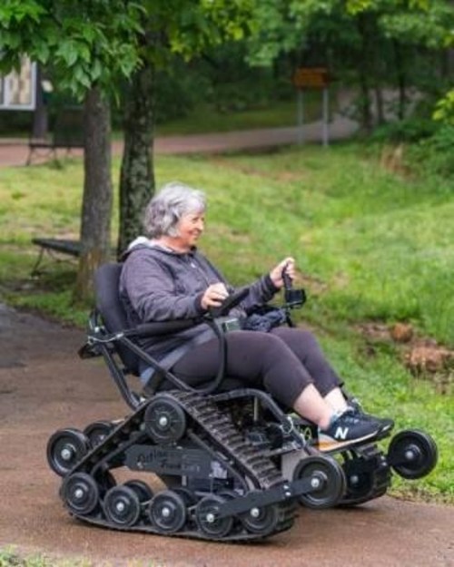 A woman in a motorized wheelchair with tracks navigates a park path surrounded by greenery.