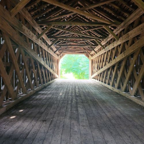 A view inside a wooden covered bridge, showcasing its beams and a bright opening at the end leading to greenery.
