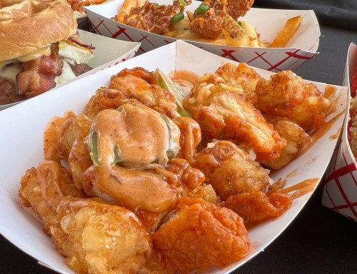 A close-up of crispy fried shrimp and tater tots drizzled with a spicy sauce, served in a paper tray.