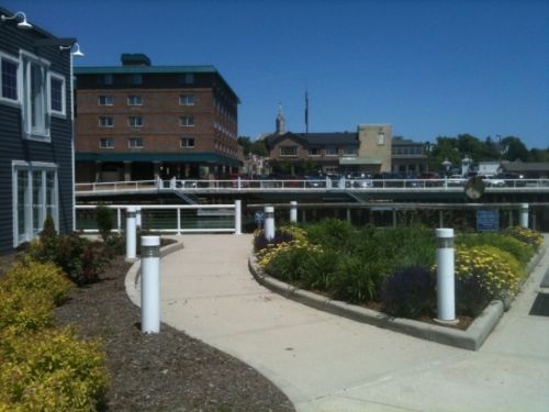 A scenic view of a waterfront area with a pathway, flowers, and buildings in the background under a clear blue sky.