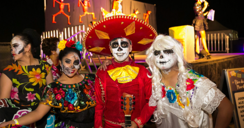 Three individuals in colorful costumes and face paint celebrate, with a festive backdrop and performers in the background.