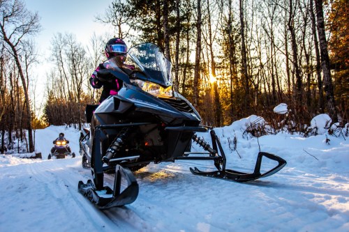 A person rides a snowmobile on a snowy trail, surrounded by trees and a sunset in the background.