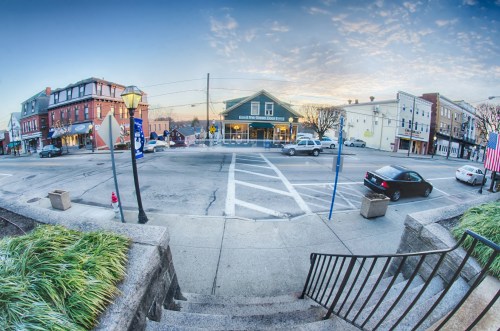 A fisheye view of a street corner with shops, a car, and a lamppost under a colorful sky at dawn.