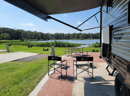 A campsite with a trailer, two chairs, and a view of a lake surrounded by greenery under a clear blue sky.