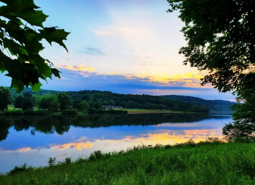 A serene lake at sunset, reflecting colorful clouds and surrounded by lush greenery and trees.