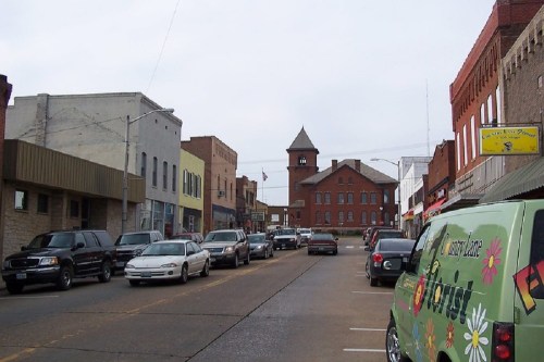 A small town street lined with parked cars, featuring brick buildings and a clock tower in the background.