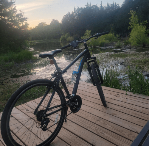 A bicycle rests on a wooden deck by a serene pond surrounded by greenery at sunset.