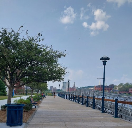 A scenic waterfront walkway lined with trees, benches, and lampposts under a partly cloudy sky.