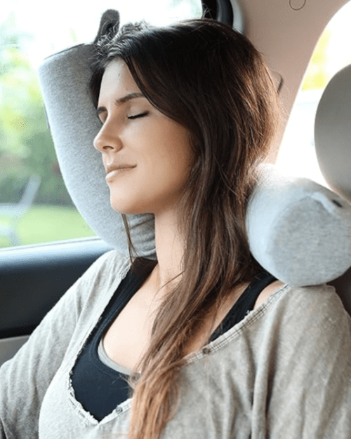 A woman with long hair relaxes in a car, eyes closed, resting her head on a gray neck pillow.
