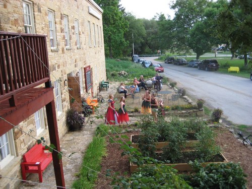 A group of people in colorful dresses dance outside a stone building, with gardens and parked cars nearby.