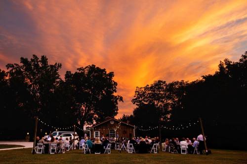 A gathering of people seated outdoors under a vibrant sunset, with string lights illuminating a rustic venue.