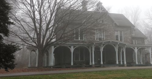 A foggy scene of a large, historic stone house with a wraparound porch and bare trees in the foreground.