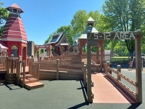Colorful playground with wooden structures and ramps, labeled "5-12 AREA," surrounded by green trees under a clear blue sky.