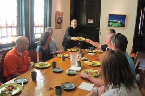 A group of people seated at a table enjoying a meal, with one person serving food to another. Colorful stained glass in the background.