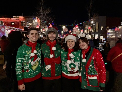 Four friends in festive holiday sweaters pose together at a nighttime outdoor event, surrounded by colorful lights.