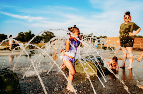 Children play in a splash pad, running through water jets while an adult watches nearby on a sunny day.