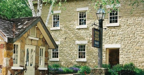 A charming stone building with a small cottage and a sign, surrounded by greenery and flowers.