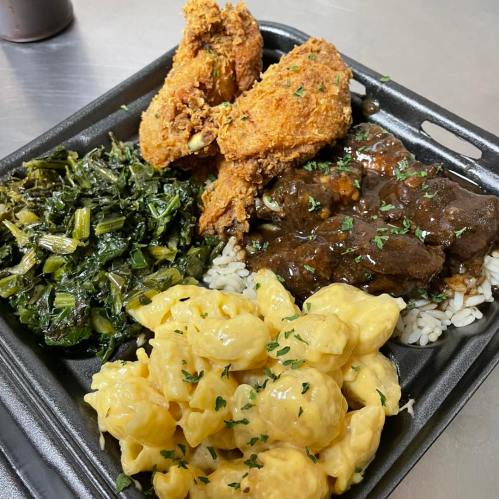 A takeout container with fried chicken, collard greens, beef stew over rice, and macaroni and cheese, garnished with parsley.