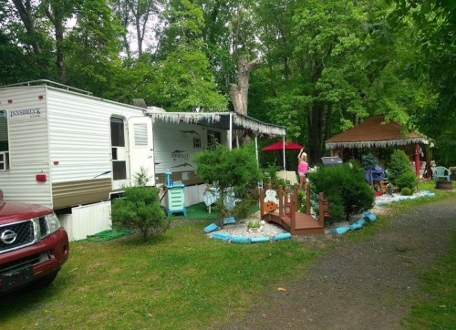 A camper surrounded by greenery, with a small deck, decorations, and a child playing nearby.