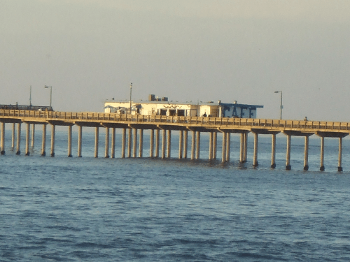 A wooden pier extends over calm water, leading to a white building with blue accents in the background.