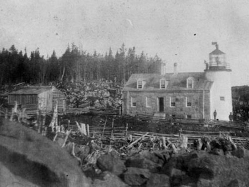Black and white photo of a lighthouse and keeper's house surrounded by trees and rocky terrain.
