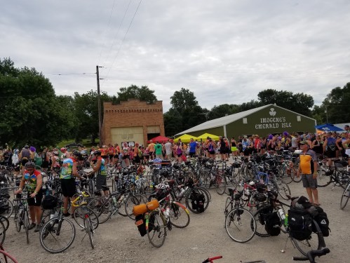 A large crowd of cyclists gathers near a bike parking area, with tents and a building in the background.