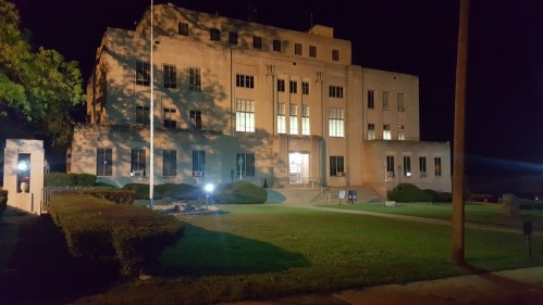 A large, illuminated building at night, surrounded by well-maintained lawns and trees.