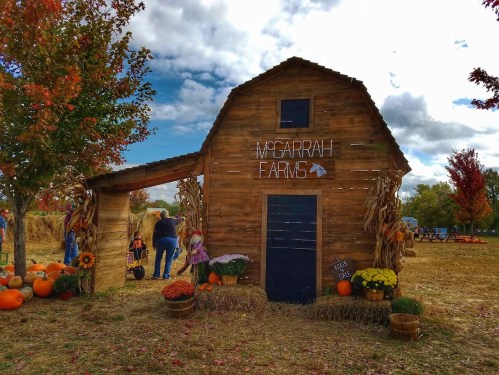 A rustic wooden barn with "McGarrah Farms" sign, surrounded by pumpkins and colorful autumn foliage.