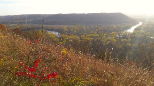 A scenic view of a river winding through lush, colorful autumn foliage and rolling hills under a bright sky.