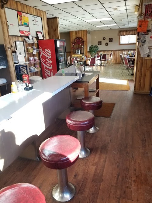 A cozy diner interior featuring a counter with red stools, wooden paneling, and a Coca-Cola display.