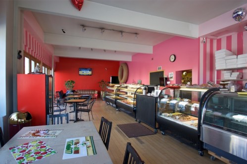 Bright café interior with pink walls, display cases of pastries, and seating. A clock and TV are visible on the wall.