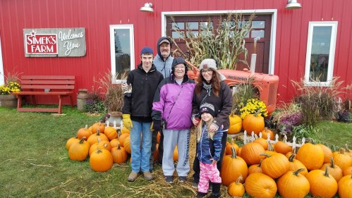 A family poses in front of a red barn with pumpkins, smiling together on a fall day at a farm.