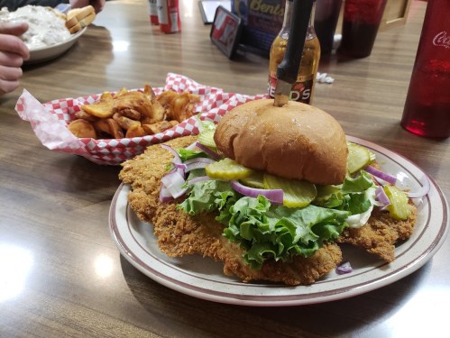A large fried chicken sandwich with lettuce, pickles, and onions, served with a side of potato wedges.