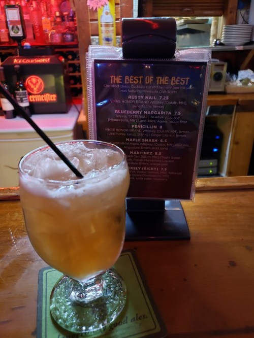 A cocktail glass filled with a drink sits on a bar counter next to a menu featuring various cocktails.