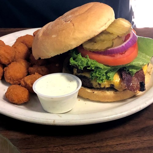 A plate with a burger topped with lettuce, tomato, pickles, and bacon, alongside a serving of crispy fried bites and dipping sauce.