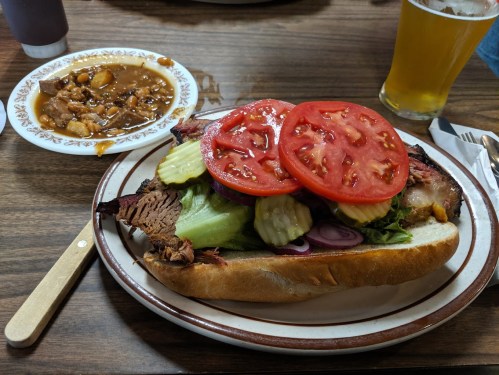 A large sandwich with brisket, lettuce, tomatoes, pickles, and onions, served with a side of baked beans and a beer.