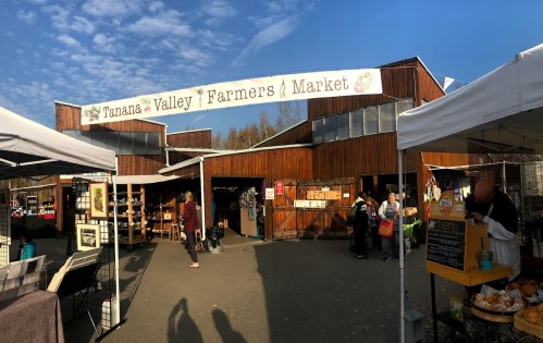 Entrance to the Tanana Valley Farmers Market, featuring wooden buildings and market stalls under a blue sky.