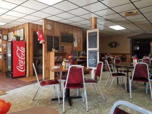 A cozy diner interior with red chairs, wooden paneling, a Coca-Cola vending machine, and chalkboard menus.