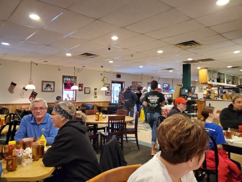 A busy diner scene with patrons seated at tables, chatting, and staff serving in a cozy, casual atmosphere.