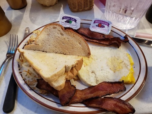 A plate with two slices of toast, crispy bacon, hash browns, and a fried egg, accompanied by jelly packets.